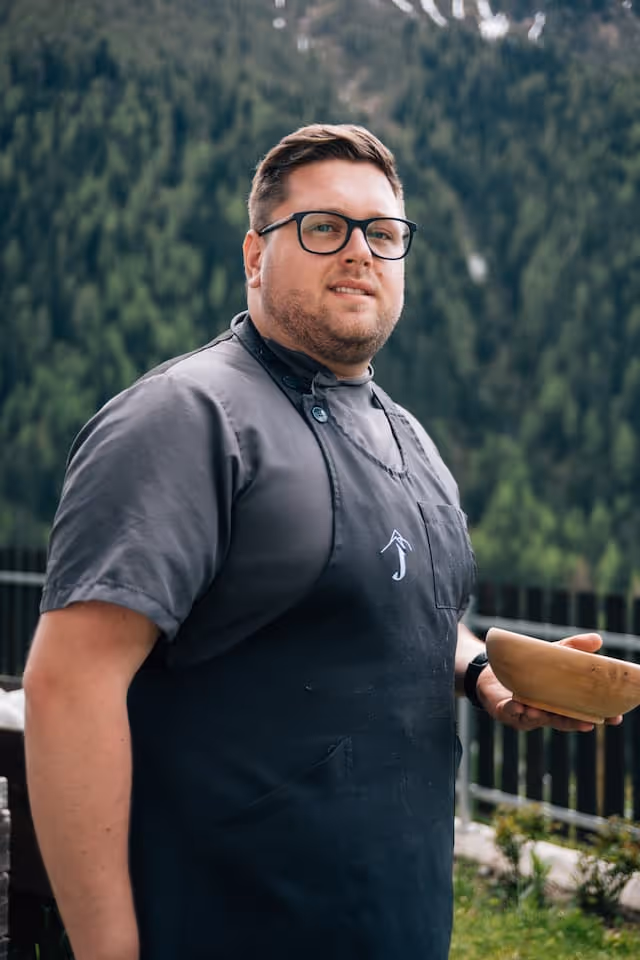 Porträt eines Mannes mit Brille, der eine schwarze Schürze trägt und eine braune Schüssel hält, mit bewaldeten Bergen im Hintergrund.