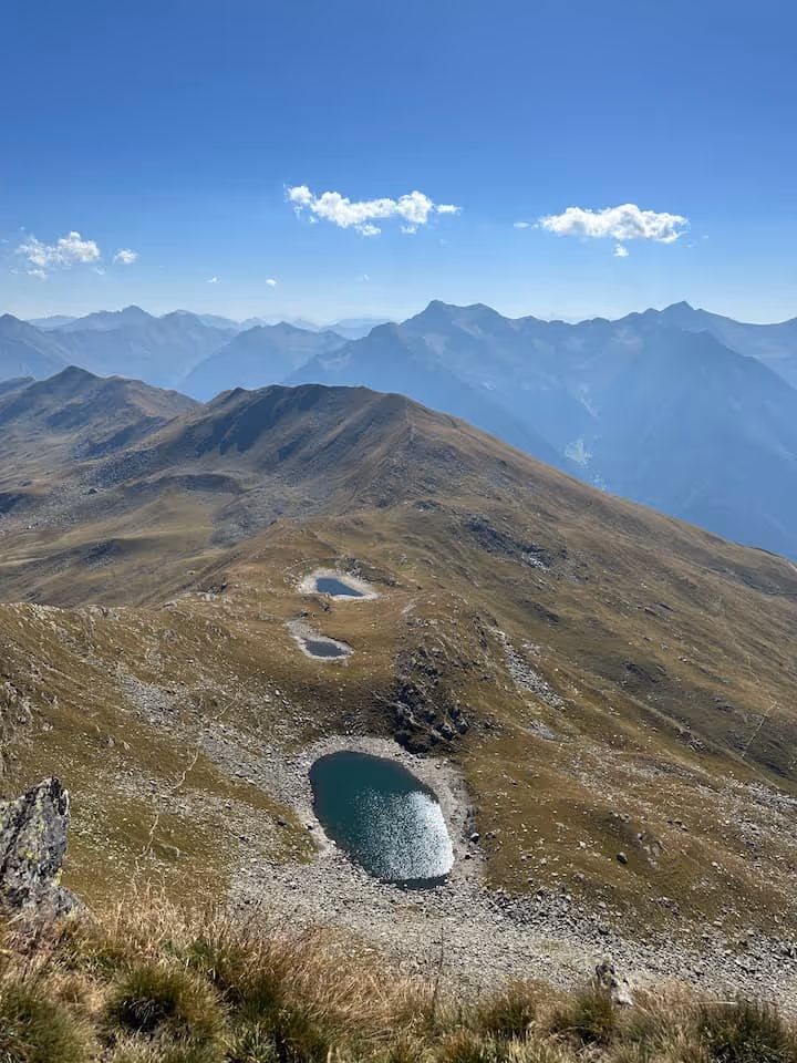 Blick auf drei Bergseen auf einem grasbewachsenen Bergrücken mit dahinterliegenden nebligen Bergketten unter blauem Himmel.