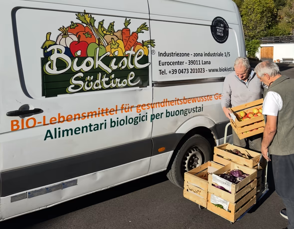 Zwei Männer laden Holzkisten mit frischem Bio-Obst und Gemüse aus einem Lieferwagen mit Biokiste Südtirol Beschriftung.