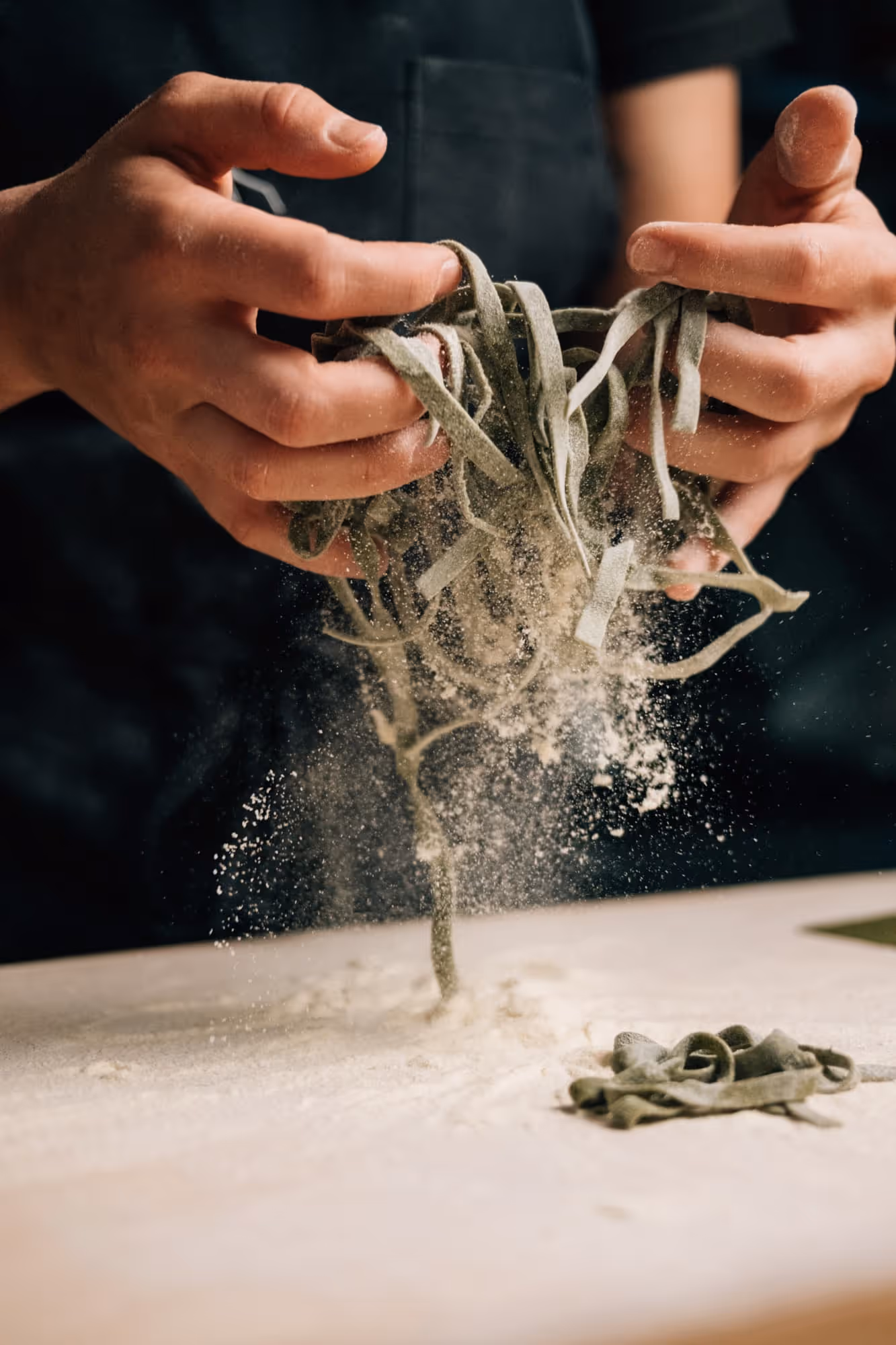 Hände, die frische grüne Tagliatelle über einer mit Mehl bestäubten Holzfläche halten.
