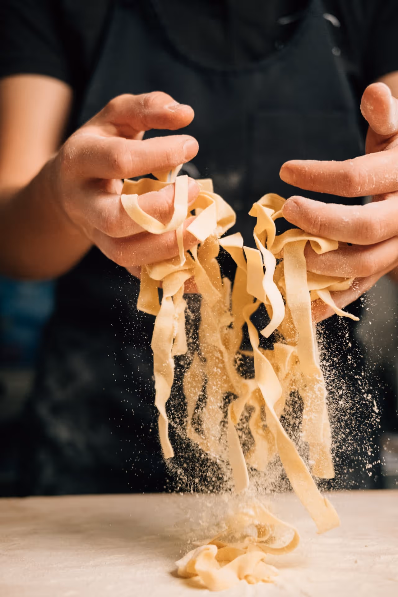 Hände, die frische Bandnudeln aus Teig über eine bemehlte Oberfläche halten und leicht fallen lassen.