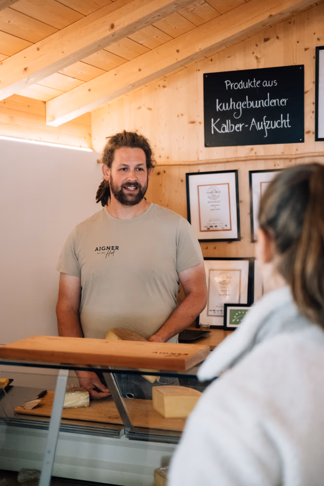 Verkäufer hinter einer Holztheke mit Käse vor sich, Gespräch mit einer Kundin, im Hintergrund Schilder und Zertifikate an Holzwand.
