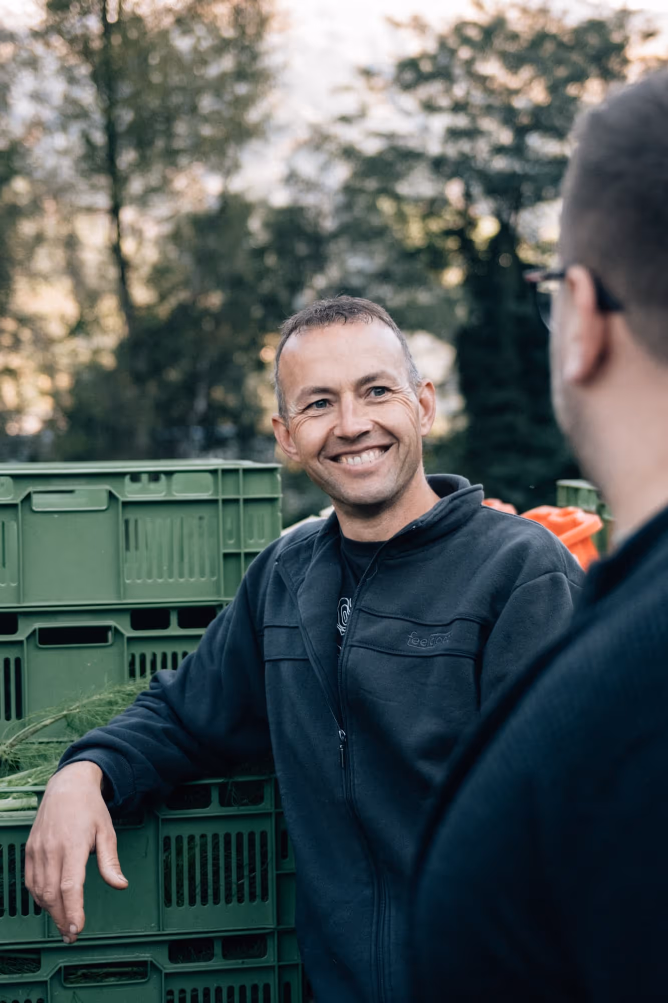 Fröhlicher Mann lehnt an grünen Plastikbehältern und unterhält sich mit einem anderen Mann im Freien.