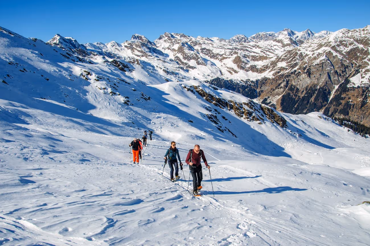 Vier Personen beim Schneeschuhtrekking in verschneiter Berglandschaft mit klar blauem Himmel und schroffen Gipfeln im Hintergrund.
