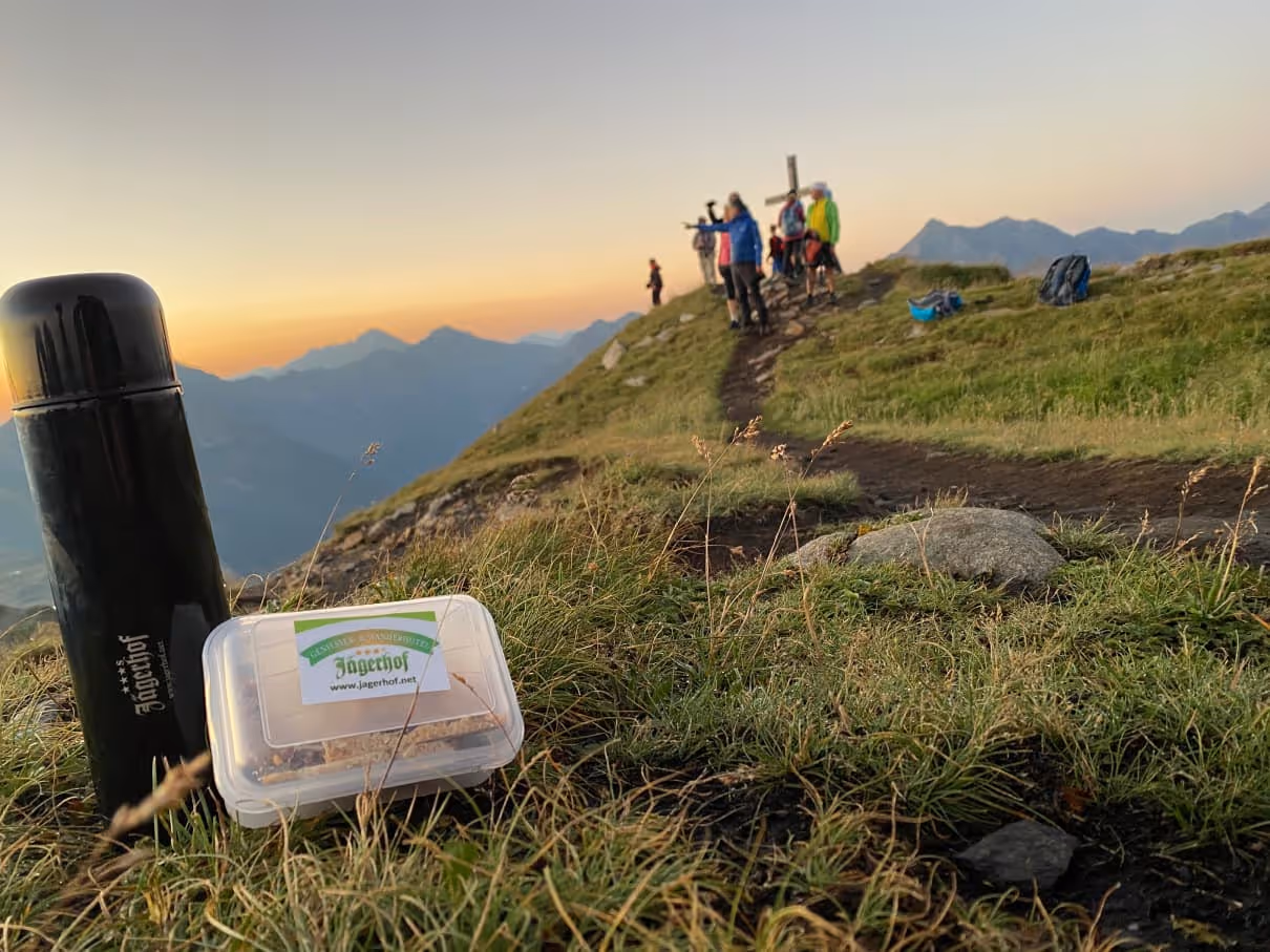 Thermosflasche und Snackbox auf Gras mit Wanderern auf einem Berggipfel und Sonnenuntergang im Hintergrund.