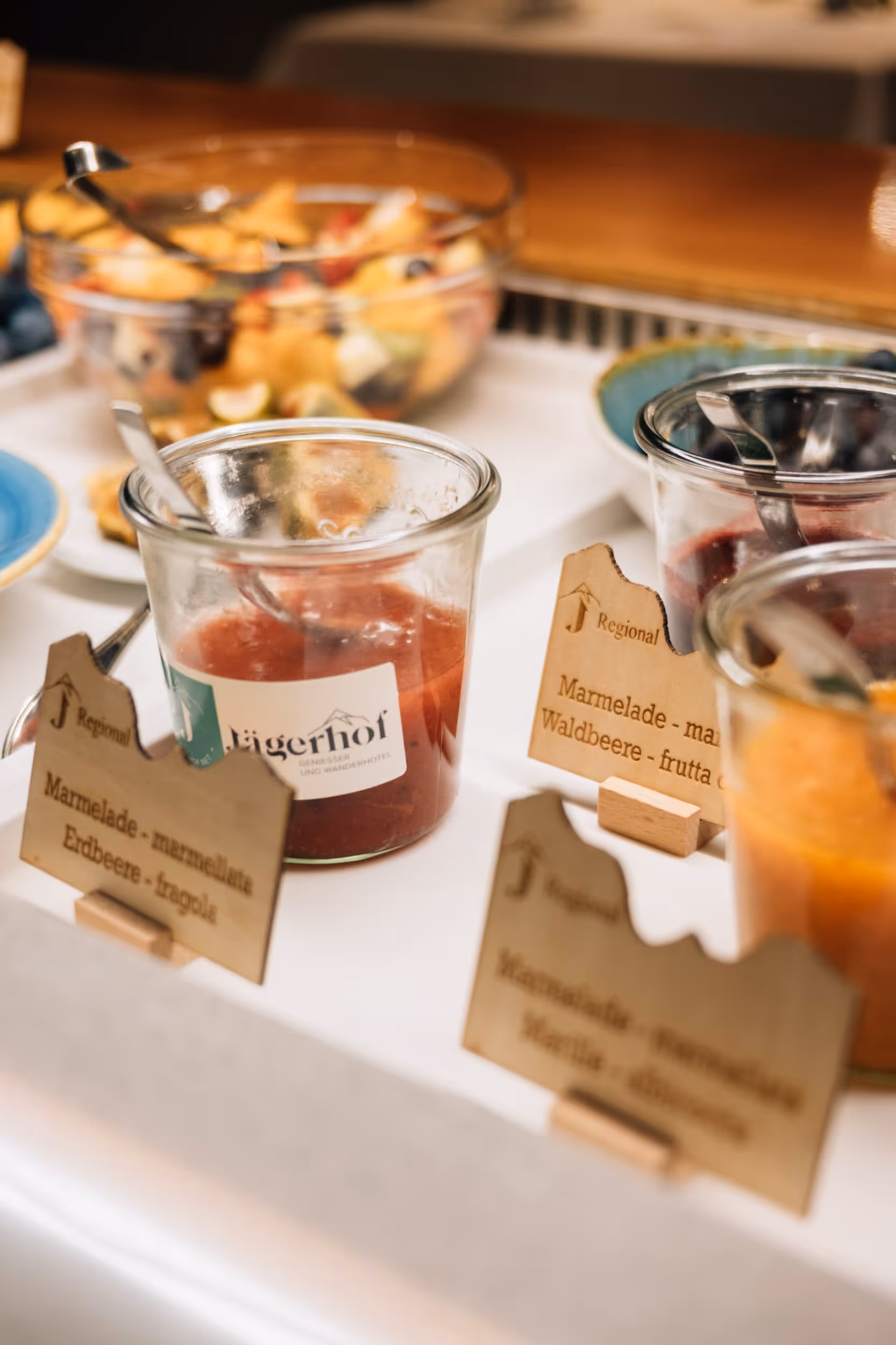 Gläser mit verschiedenen Marmeladen auf einem Frühstücksbuffet, im Hintergrund eine Schüssel mit Obstsalat.