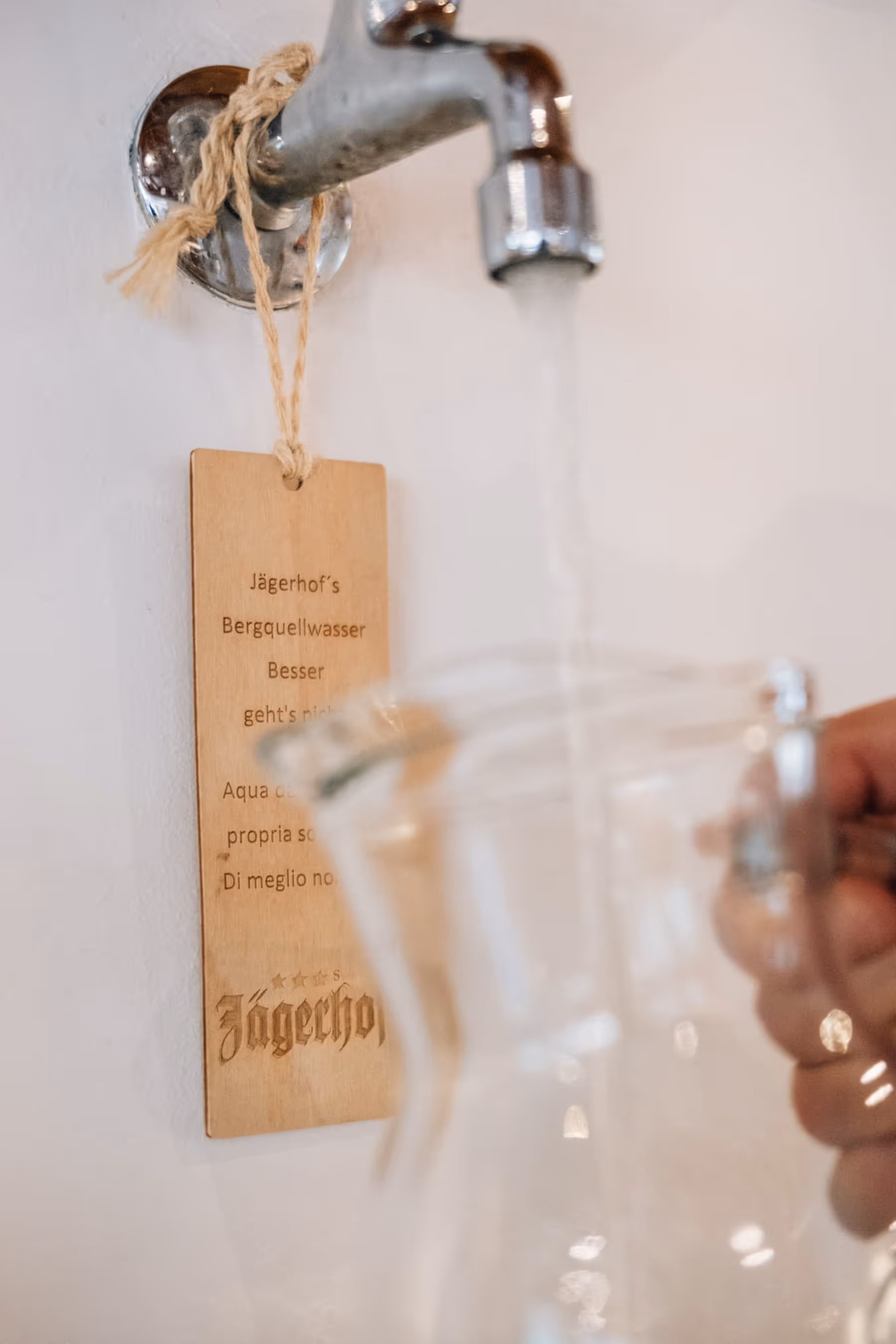 Hand füllt Glaskrug mit Wasser aus einem Wasserhahn, an dem ein Schild mit dem Text „Jägerhof’s Bergquellwasser Besser geht's nicht“ hängt.
