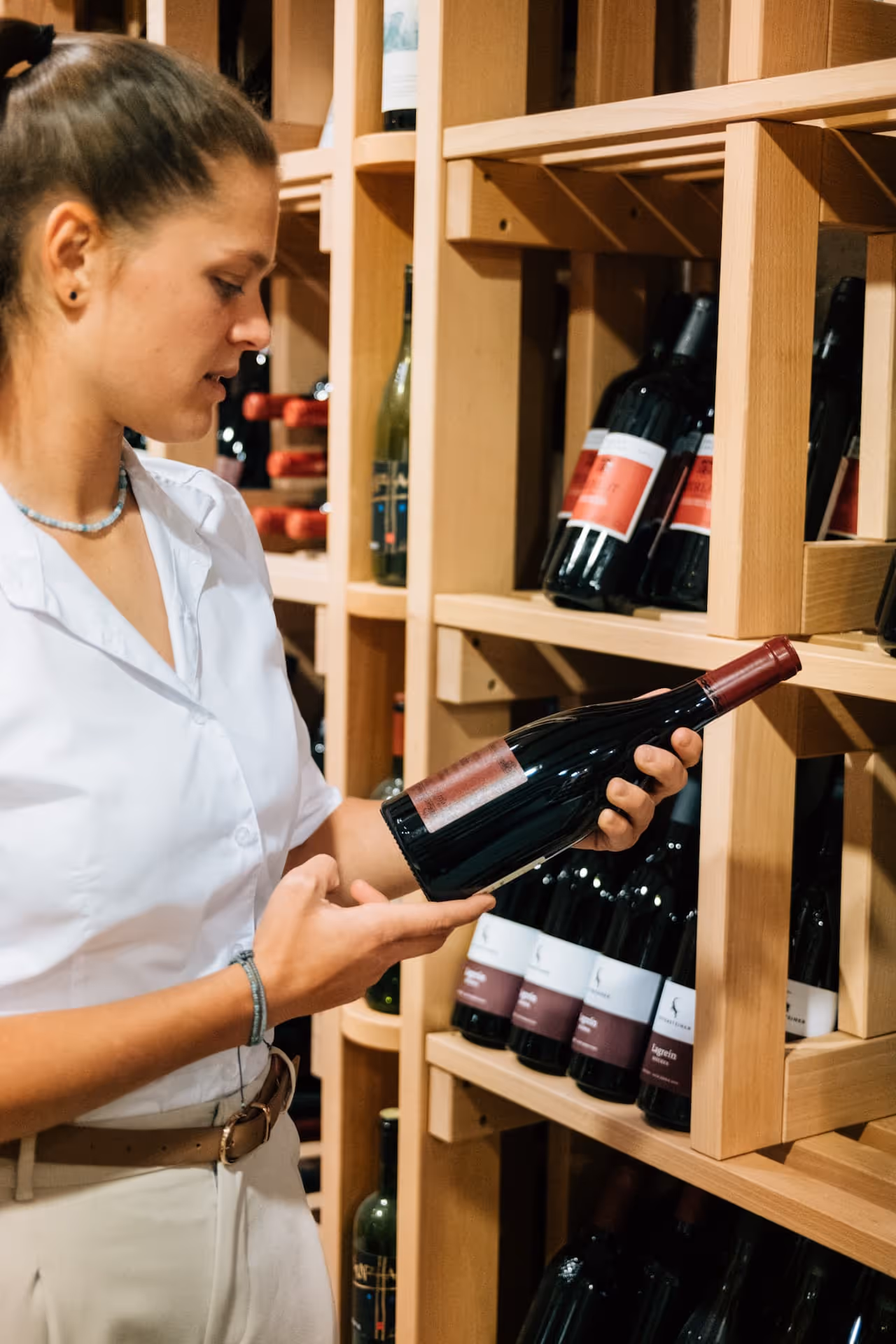 Frau in weißem Hemd hält und betrachtet eine Flasche Rotwein vor einem Weinregal aus Holz.