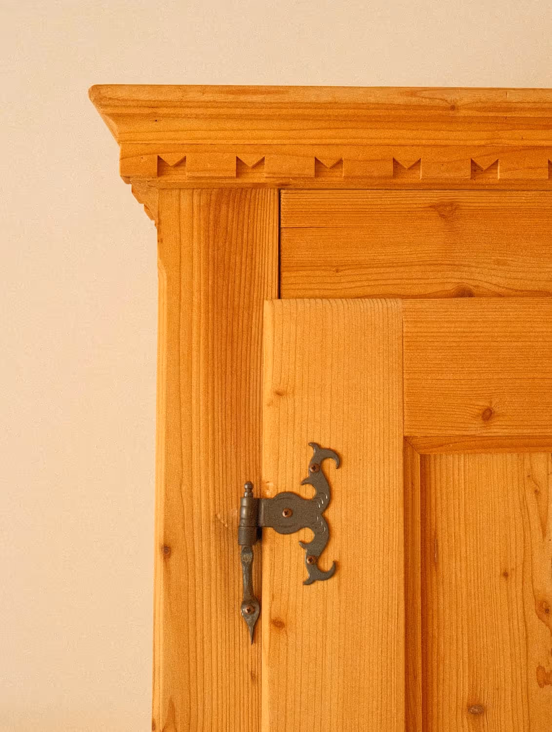 Nahaufnahme eines Schmuck-Türscharnier aus schwarzem Metall an einer hellen Holztür mit Zierleiste oben.