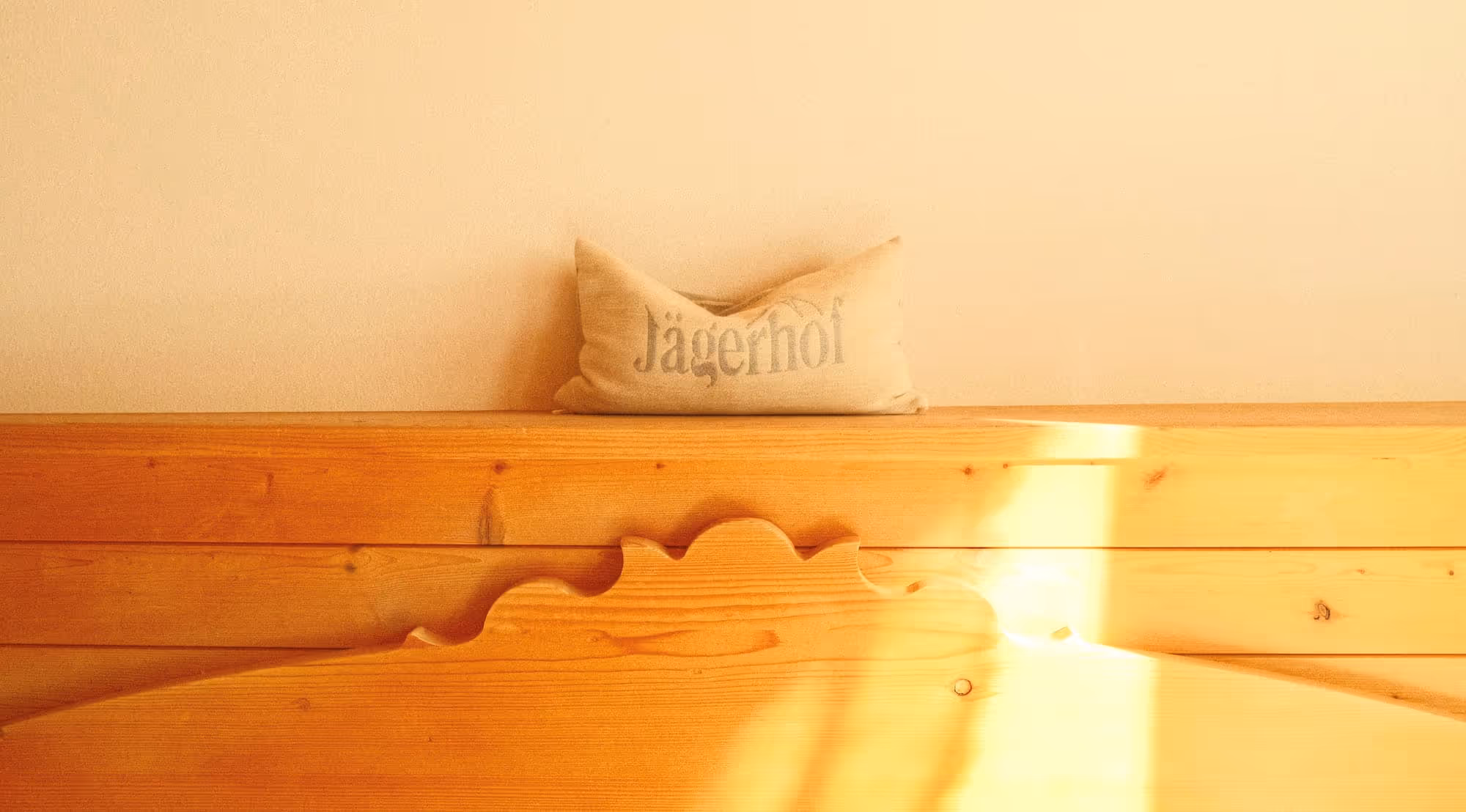 Ein beiges Kissen mit der Aufschrift ‚Jägerhof‘ auf einer hölzernen, geschnitzten Bank vor einer hellen Wand.