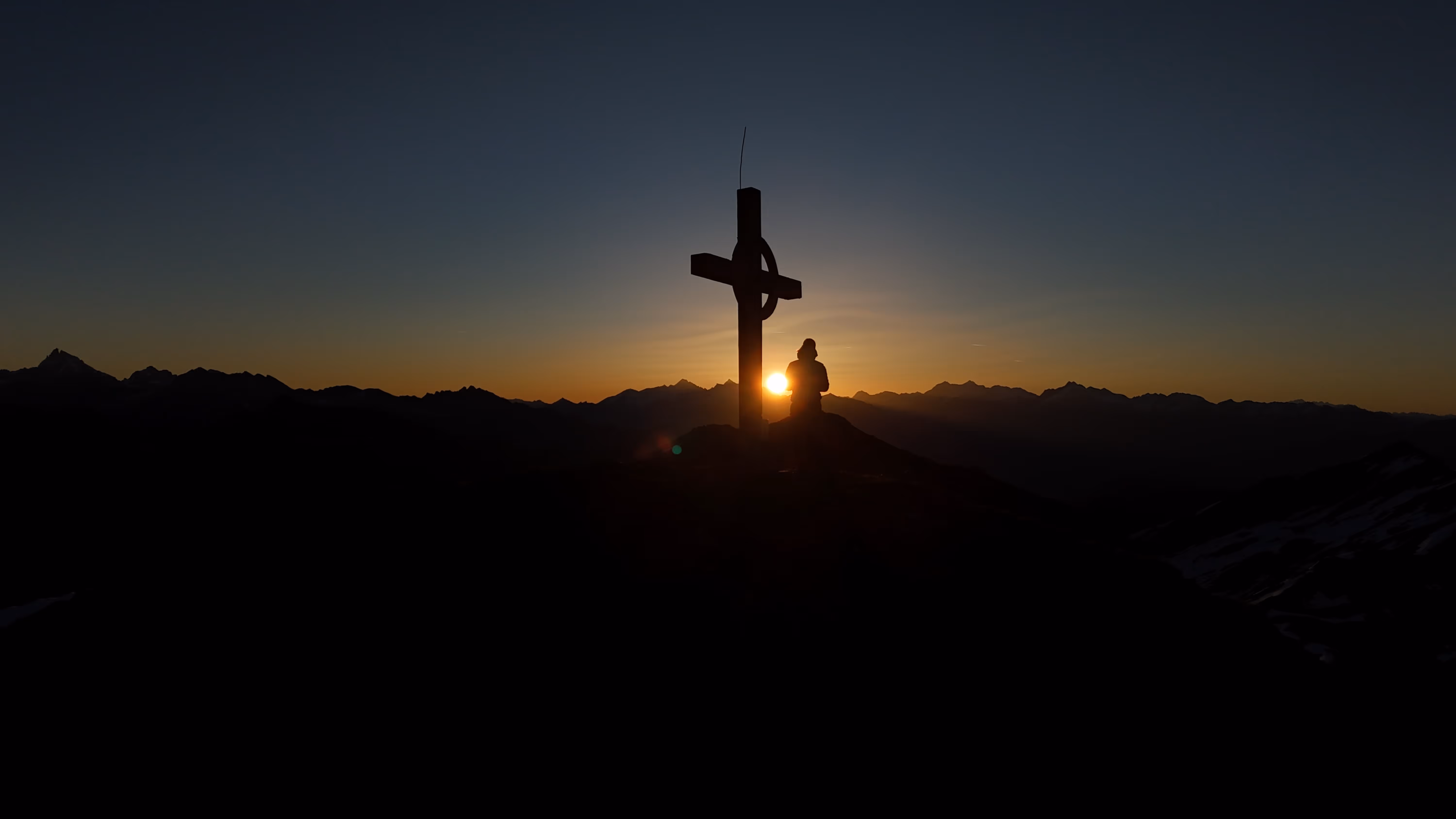Sonnenuntergang hinter einem Gipfelkreuz mit einer Person, die vor den Bergen steht.