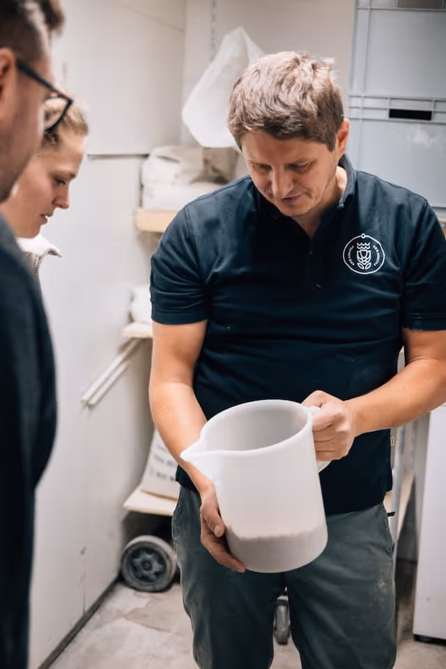 Mann in schwarzem Hemd zeigt einen weißen Messbecher mit Mehl vor zwei anderen Personen in einem Raum mit Regalen und Mehlsäcken.