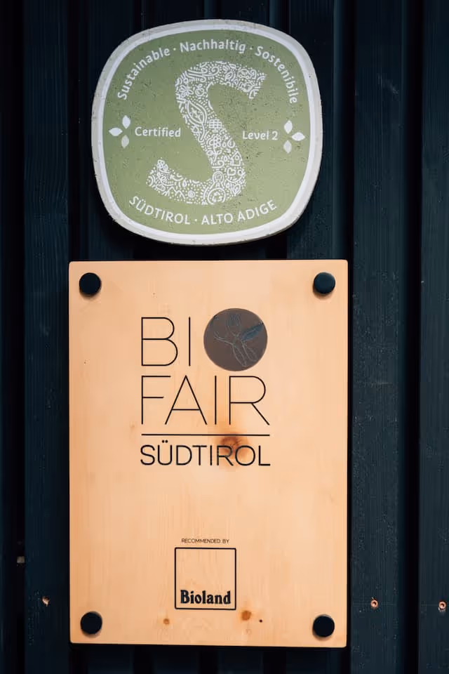 Zwei Schilder an einer dunklen Wand mit Nachhaltigkeitszertifikaten, oben das grüne Siegel mit dem Text Nachhaltig Stufe 2, unten Holzschild mit der Aufschrift Bio Fair Südtirol und Bioland-Empfehlung.