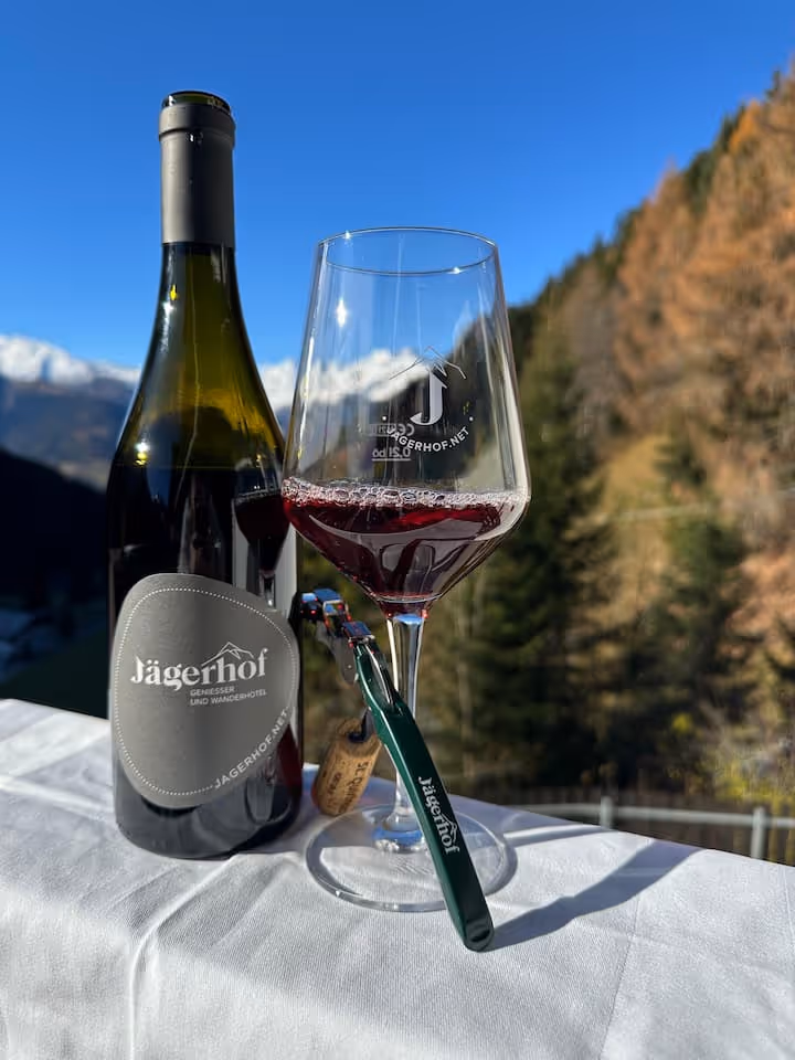 Flasche und Weinglas mit rotem Wein, beides mit Jägerhof-Logo, auf weißer Tischdecke vor Berglandschaft an sonnigem Tag.