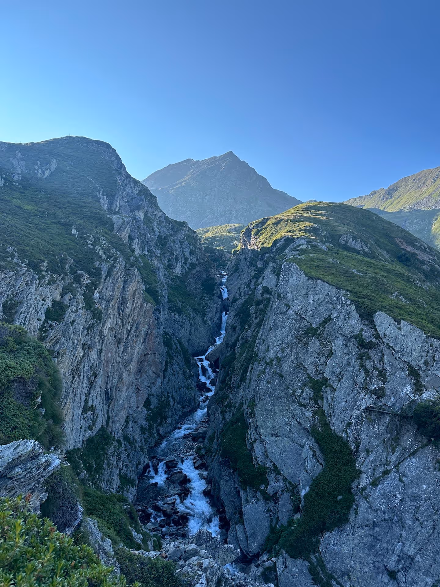 Berglandschaft mit schluchtartigem Fluss und kleinen Wasserfällen unter klarem blauem Himmel.