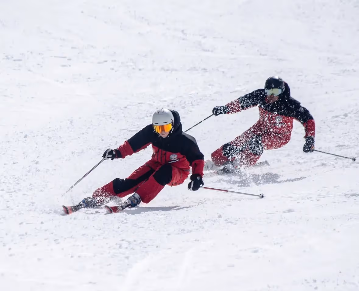 Zwei Skifahrer in rote und schwarze Skianzügen fahren schnell den schneebedeckten Hang hinunter und wirbeln Schnee auf.