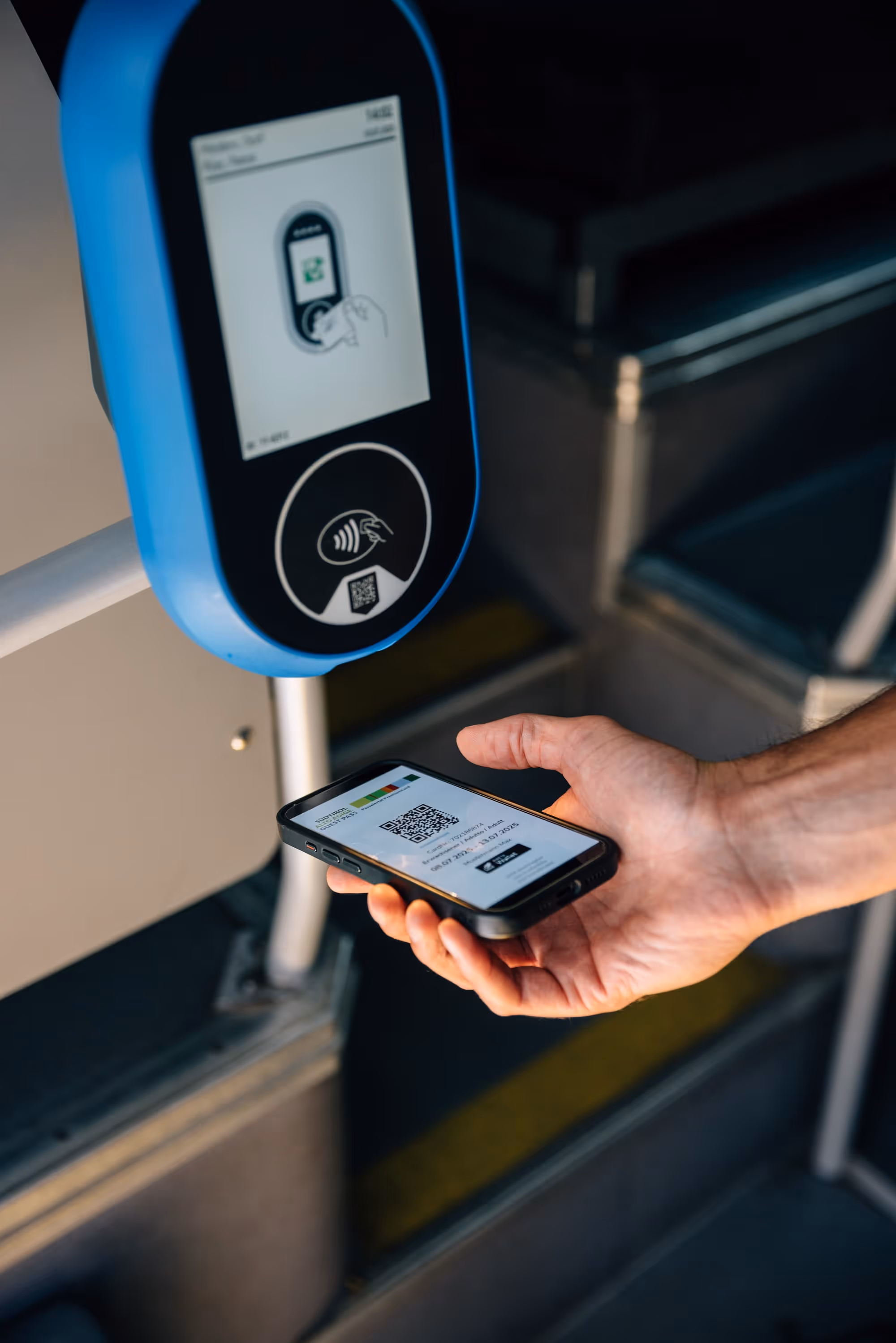 Eine Hand hält ein Smartphone mit einem QR-Code vor ein blaues kontaktloses Lesegerät zur Kontrolle.