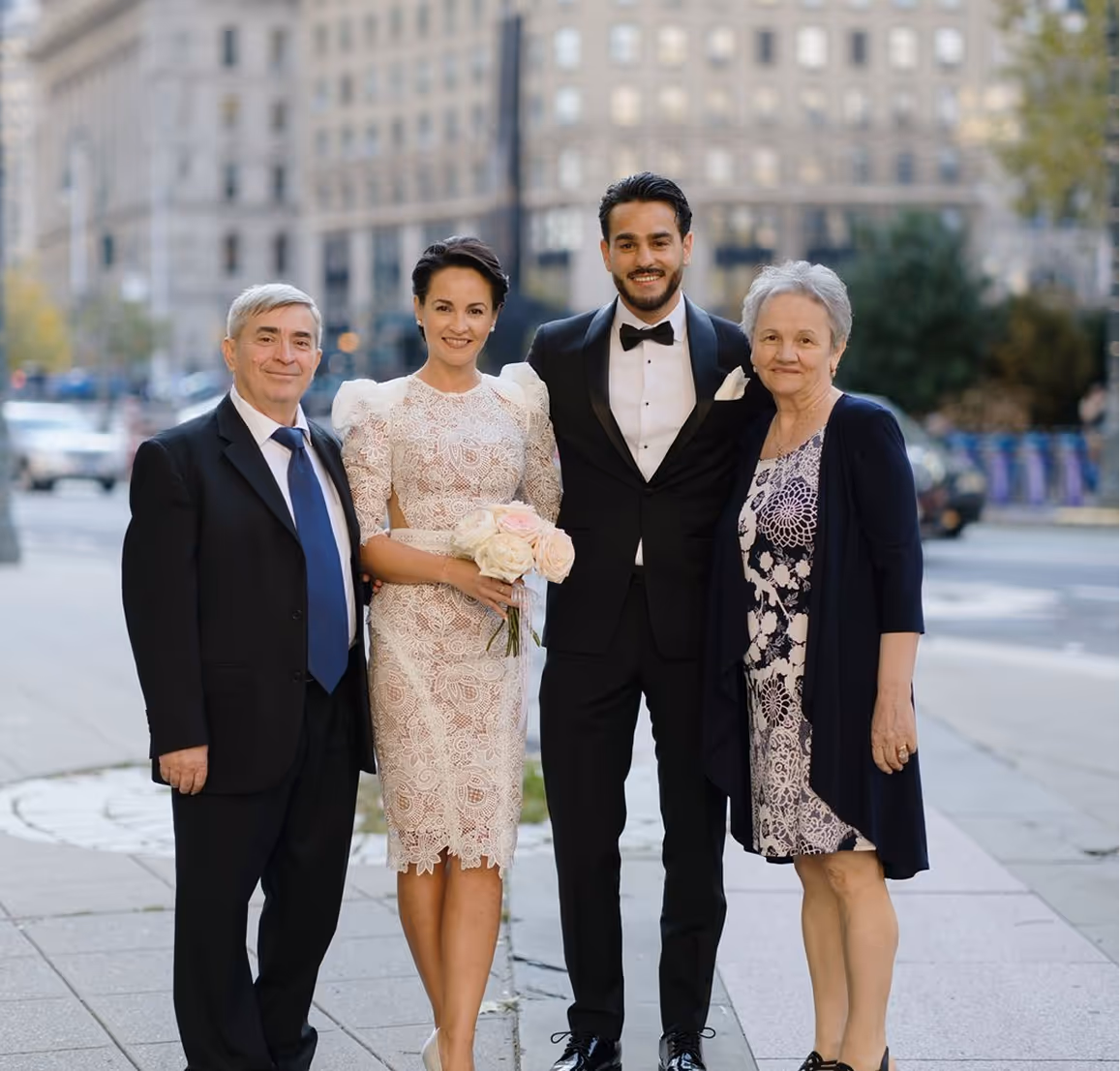 A bride in a white lace dress holding a bouquet, standing beside a groom in a black tuxedo, flanked by an older man in a suit and an older woman in a floral dress and navy cardigan on a city sidewalk.