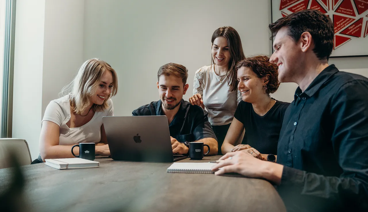 Group of young professionals gathered around a laptop, smiling and collaborating, showcasing the interactive and team-oriented approach of IT and AI (KI) training for digital commerce