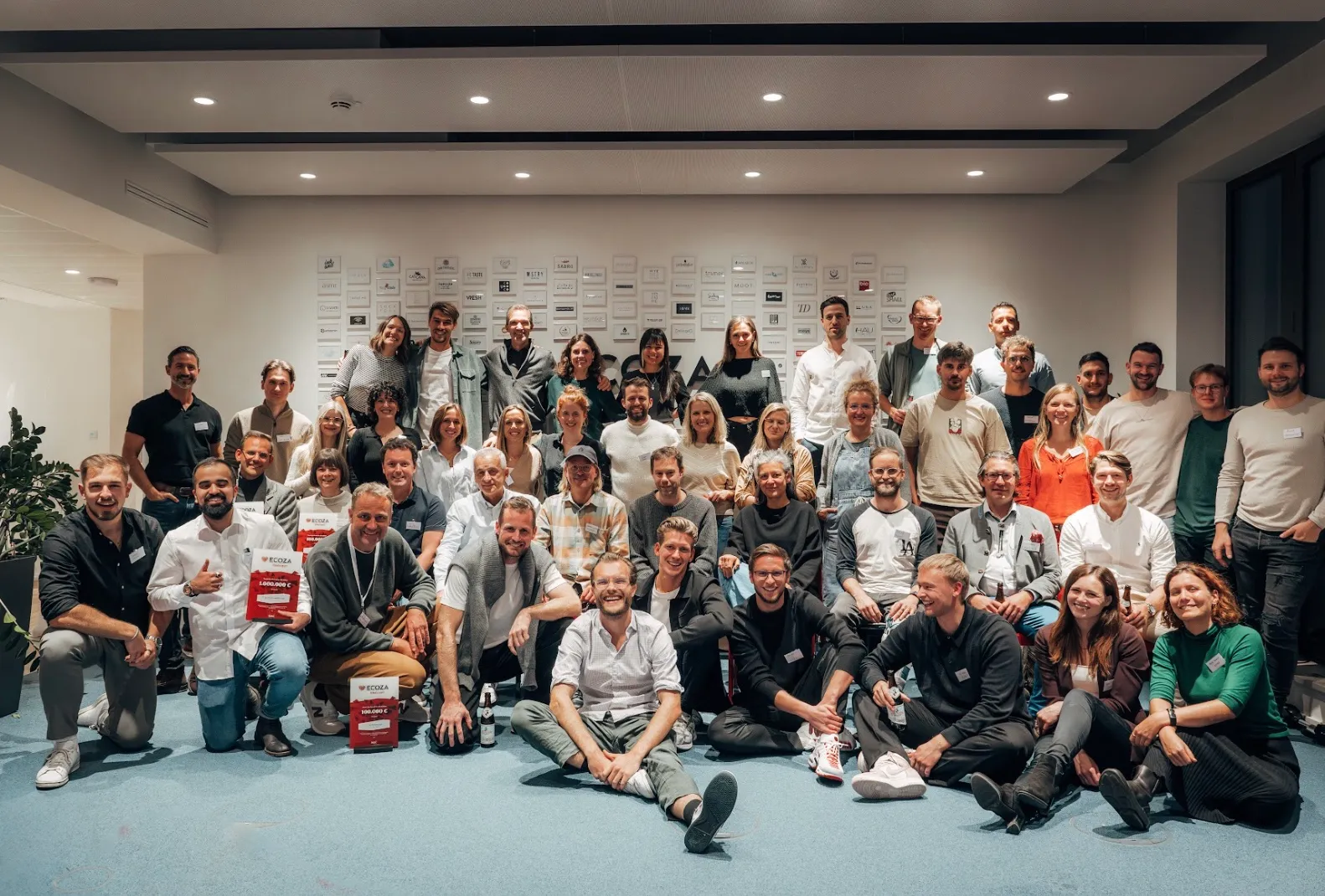 Large group photo of smiling participants and instructors gathered in front of a wall of company logos, celebrating community and collaboration in IT and AI (KI) training for digital commerce