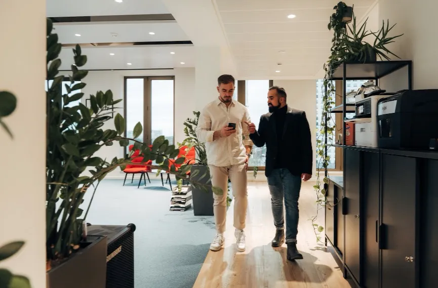 Two men walking through a bright, plant-filled office while discussing something on a phone, representing dynamic, real-world application of skills learned in IT and AI (KI) training for digital commerce