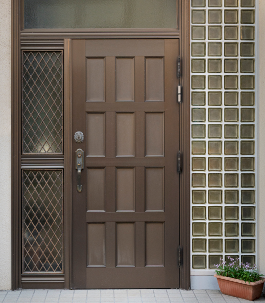 باب أمامي مغطى بألواح بنية اللون مع ألواح زجاجية مزخرفة على اليسار وكتل زجاجية على اليمين، بجوار وعاء مستطيل به زهور أرجوانية.