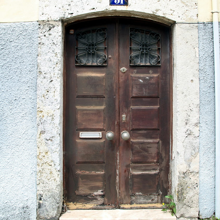 باب خشبي مزدوج بني اللون مع شبكات معدنية مزخرفة على الألواح العلوية، مثبت في إطار حجري بجدار أزرق.