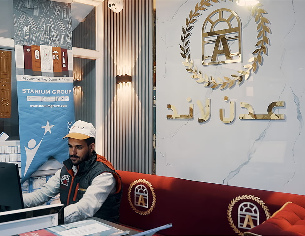 رجل يعمل على جهاز كمبيوتر في مكتب مع شعار ذهبي لشركة عدن لاند للنوافذ والأبواب على الحائط خلفه ولافتة زخرفية حول الأبواب و النوافذ ال upvc.