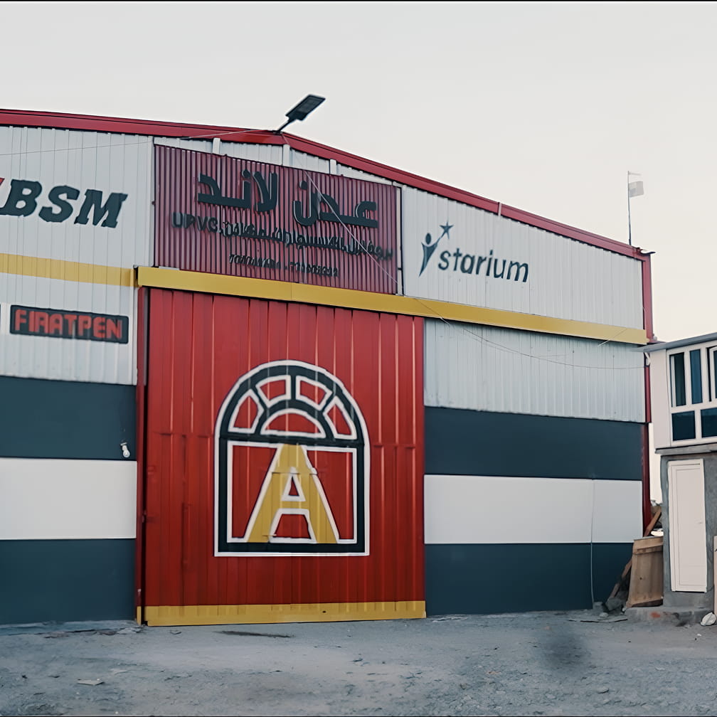منظر أمامي لمبنى صناعي بباب أحمر كبير لمصنع عدن لاند للنوافذ و الأبواب ال upvc, wpc  يحمل شعارًا على شكل نافذة يحمل الحرف A، ولافتات باللغتين العربية والإنجليزية مكتوب عليها "ستاريوم" في الأعلى.