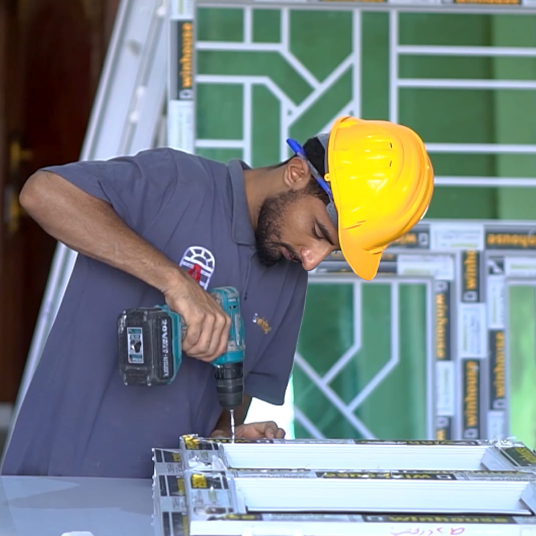 عامل يرتدي قبعة صلبة صفراء ويستخدم مثقابًا كهربائيًا لتجميع إطارات النوافذ البيضاء upvc في الداخل