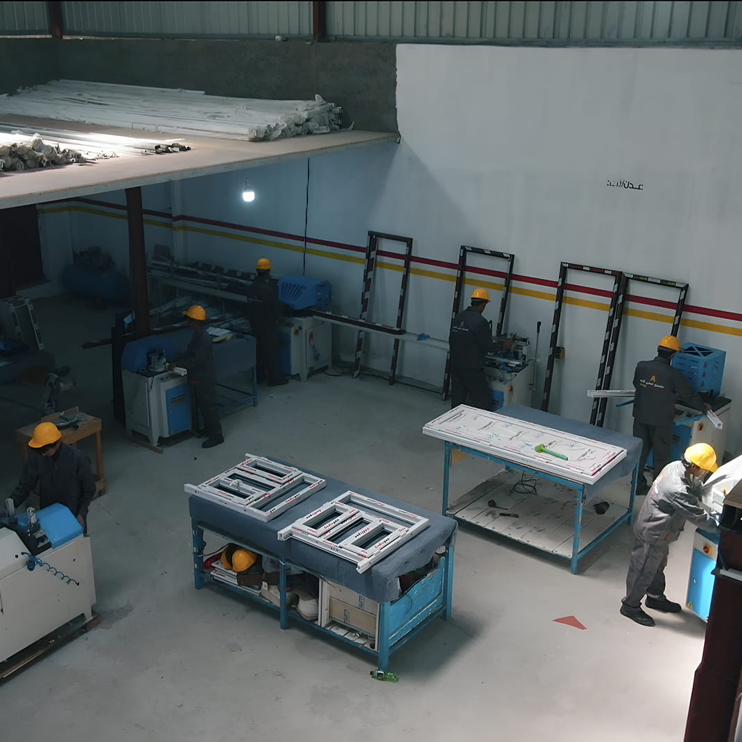 ورشة عمل مع عمال يرتدون خوذات صفراء يقومون بتجميع إطارات النوافذ على الطاولات وبالقرب من الآلات لمصنع عدن لاند للنوافذ و الأبواب ال upvc و wpc