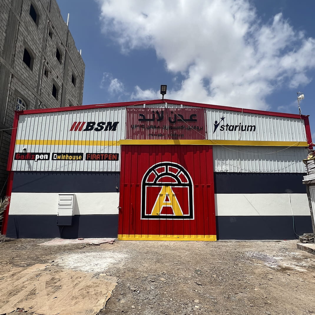 واجهة أمامية لمستودع تجاري بواجهة حمراء وبيضاء وزرقاء داكنة لمصنع عدن لاند للنوافذ و الأبواب، ويتميز بشعار كبير مع نافذة منمقة وحرف A على الأبواب الحمراء، وشعارات متعددة للعلامات التجارية أعلاه، وسماء غائمة جزئيًا في الخلفية.