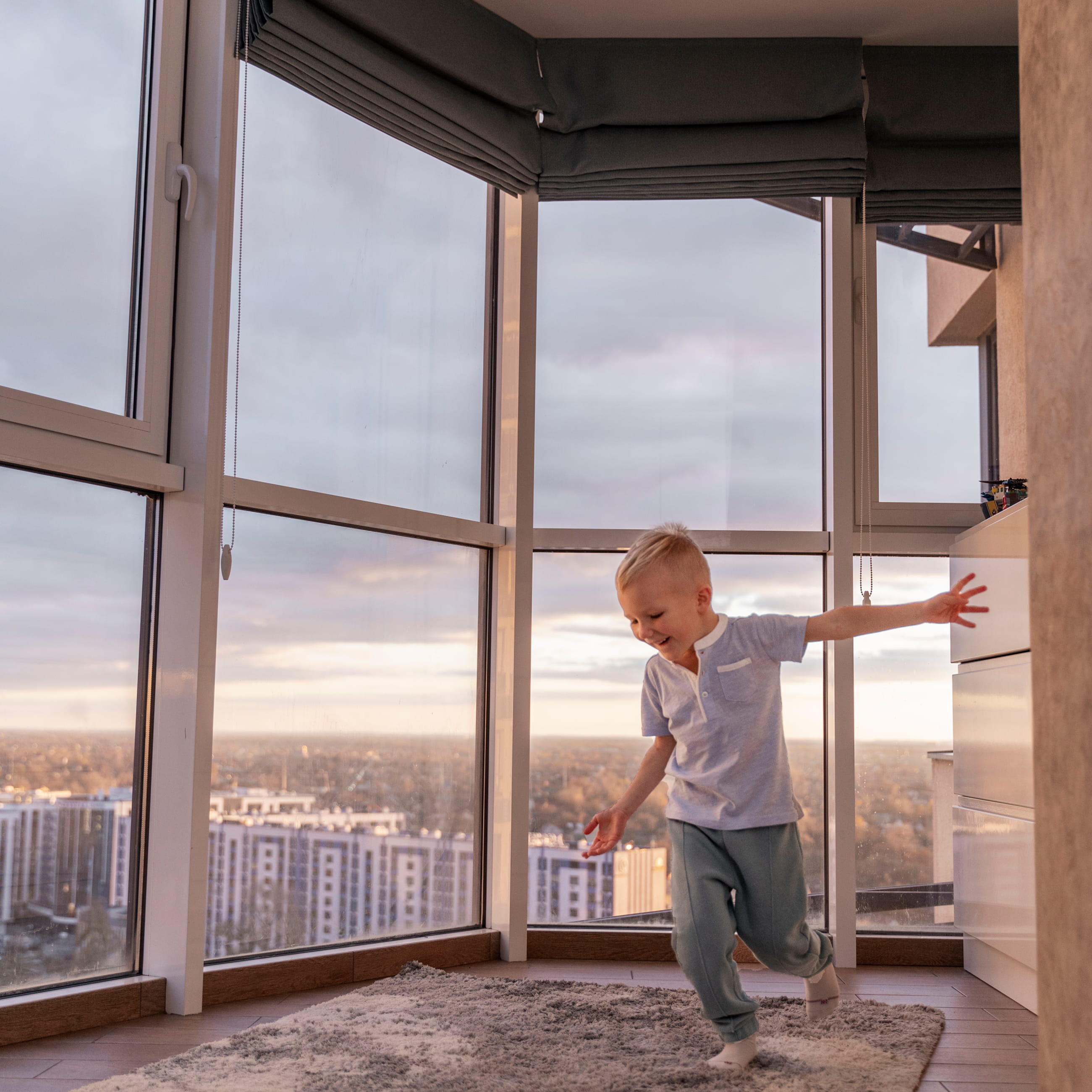 صبي صغير يتوازن بسعادة على قدم واحدة في غرفة مضاءة بنور الشمس مع نوافذ كبيرة upvc ممتدة من الأرض حتى السقف تطل على مباني المدينة عند غروب الشمس.