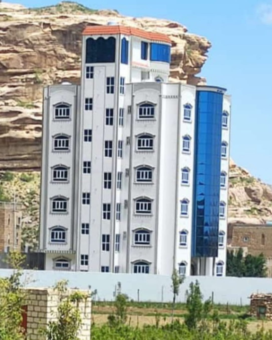 مبنى أبيض متعدد الطوابق بنوافذ عحدذ وسقف برتقالي، يقع مقابل تلال صخرية وسماء صافية.