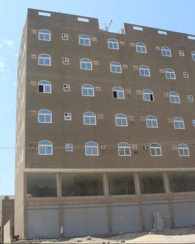Unfinished multi-story building with blue arched windows and closed metal shutters on the ground floor under a clear sky.