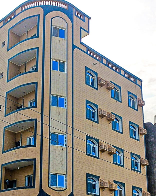 مبنى بيج مكون من أربعة طوابق مع إطارات نوافذ upvc زرقاء ووحدات تكييف متعددة وشرفات منحنية على جانب واحد.