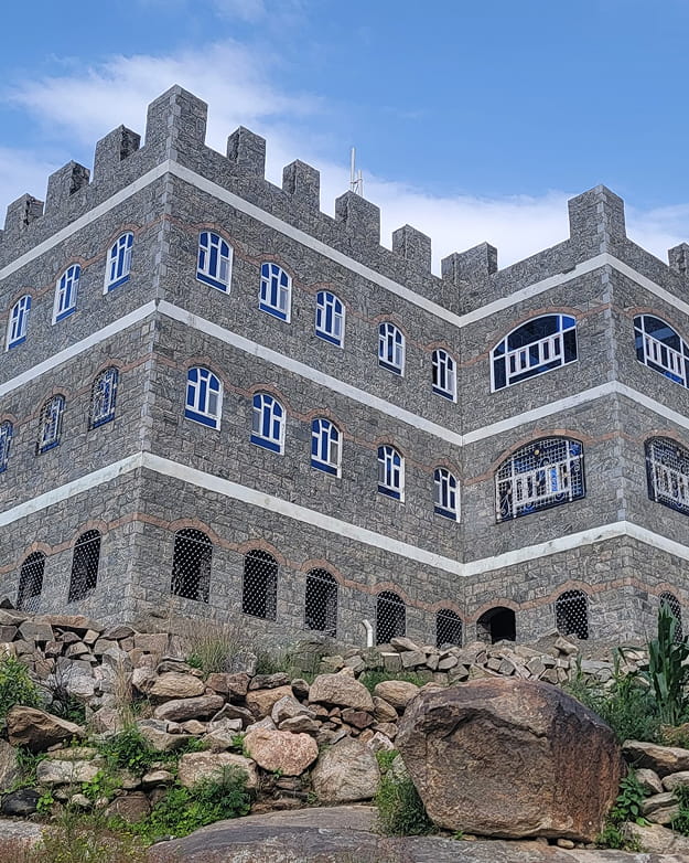 مبنى يافعي حجري يشبه القلعة بسقف مسنن ونوافذ upvc مقوسة متعددة مقابل سماء زرقاء، وتحيط به تضاريس صخرية.