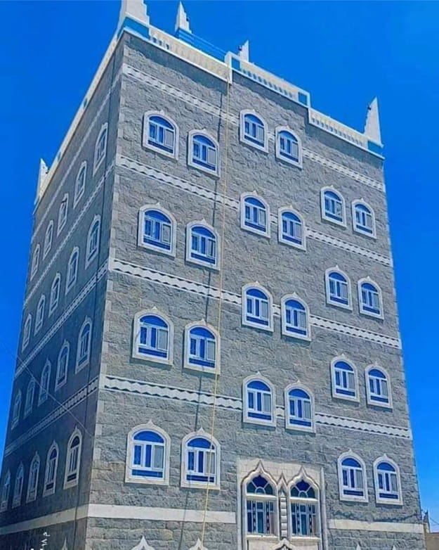مبنى يافعي حجري طويل ذو نوافذ upvc مقوسة متعددة وزخارف بيضاء مزخرفة تحت سماء زرقاء صافية.