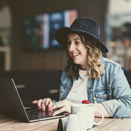Rachel, a woman in her early 30s with wavy shoulder-length hair, wearing a denim jacket and black hat, sitting in a café and smiling as she works on her laptop.