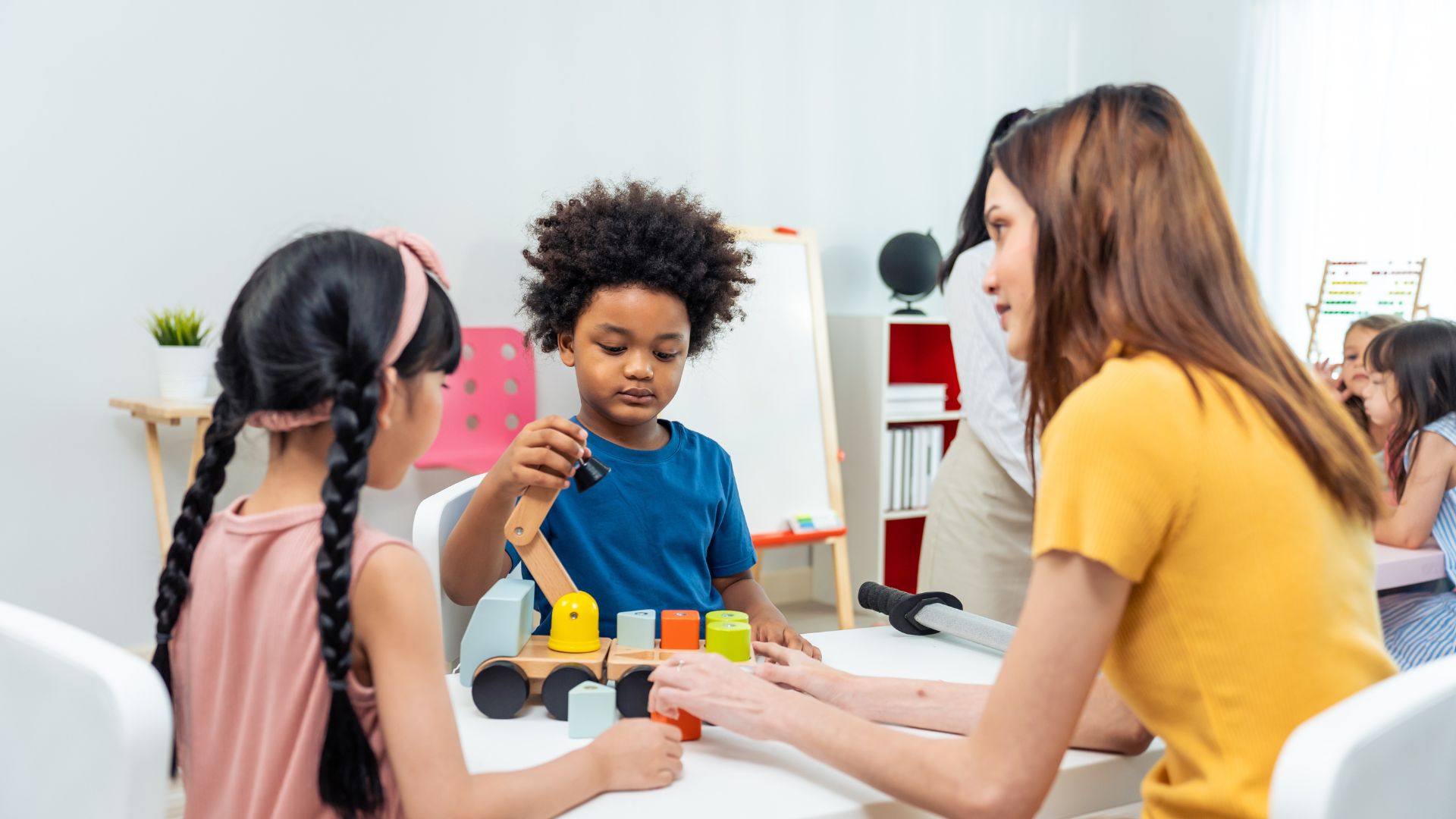 Mehrere Kinder sitzen mit einer erwachsenen Person an einem Tisch und spielen gemeinsam mit einem Holzspielzeug und bunten Bausteinen in einem hellen Raum.