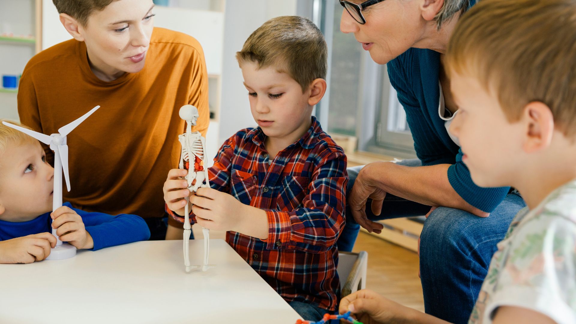 Mehrere Kinder sitzen mit zwei Erwachsenen an einem Tisch und betrachten gemeinsam ein kleines Skelettmodell