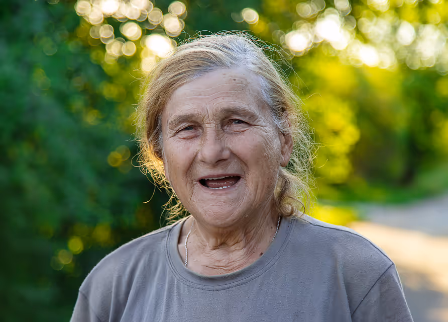 Eine ältere Frau, die lächelnd im Freien in einer grünen Umgebung steht.