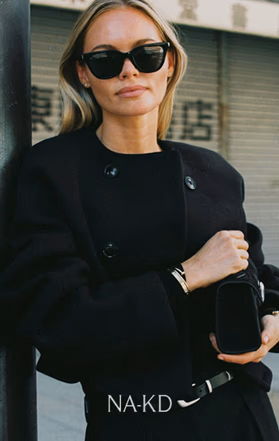 Woman wearing black sunglasses and a black coat holding a black purse, standing against a pole with a graffiti-covered wall in the background.