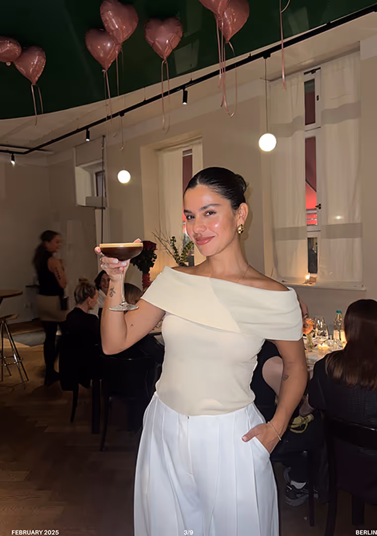 Woman in off-shoulder white top and white pants holding a cocktail glass, smiling in a warmly lit room with heart-shaped balloons.