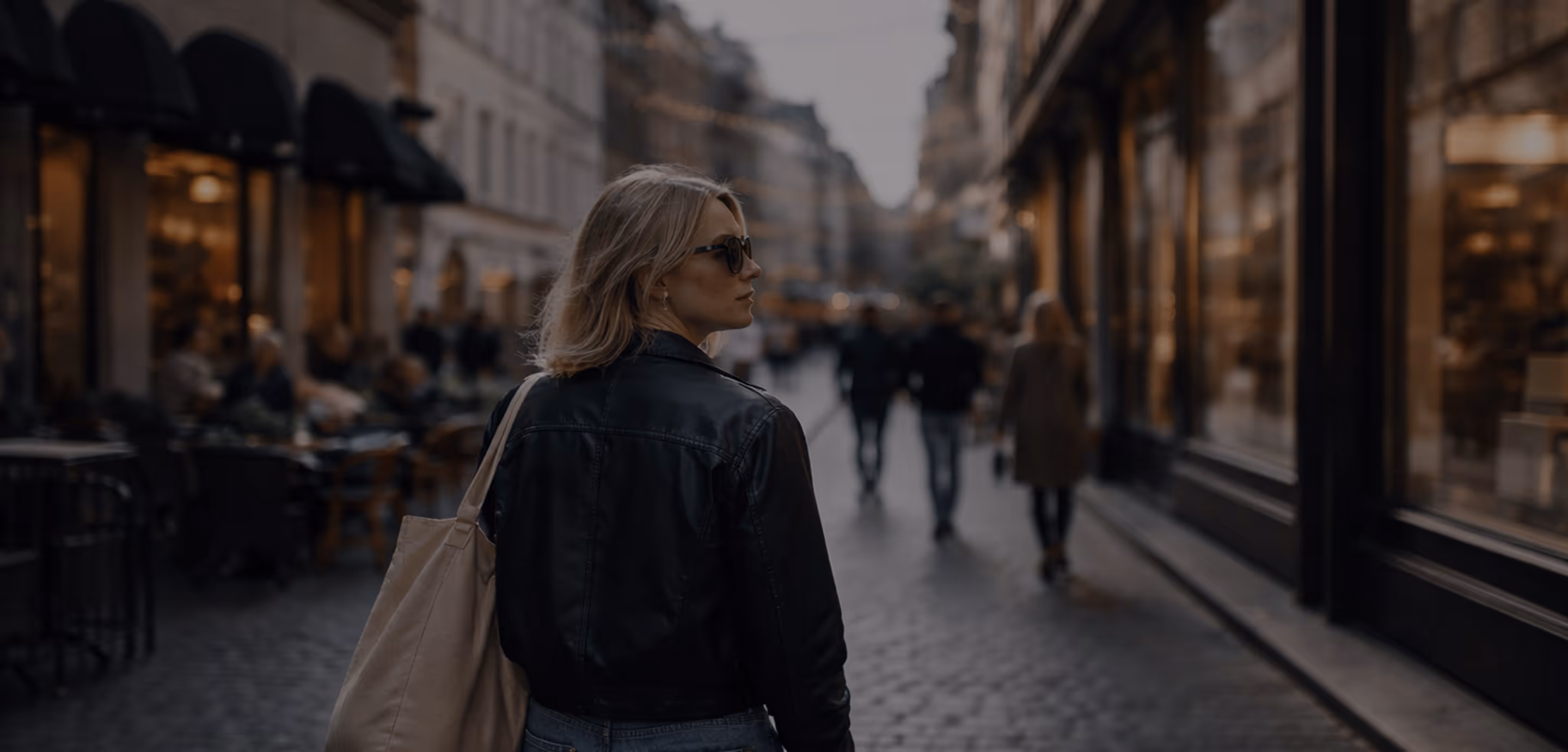 Woman in sunglasses and black leather jacket walking on a city street with shops and people in the background.