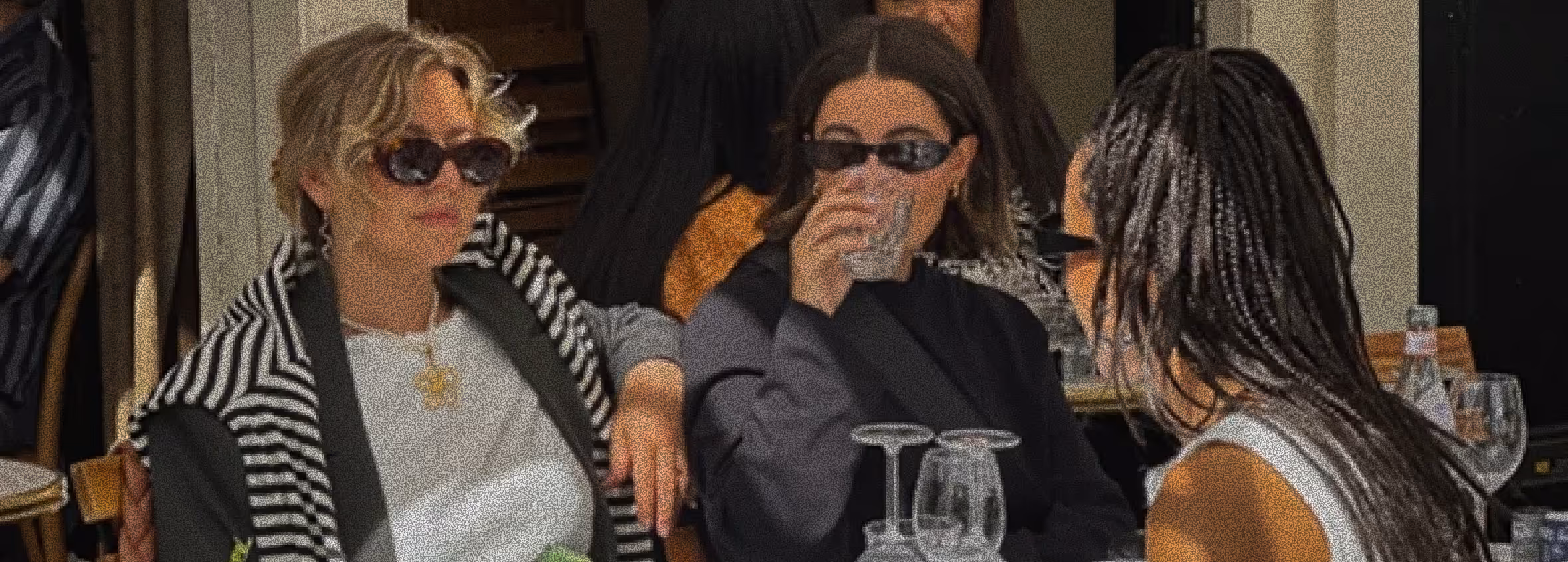 Three women wearing sunglasses seated at a table outdoors, with one drinking from a glass.