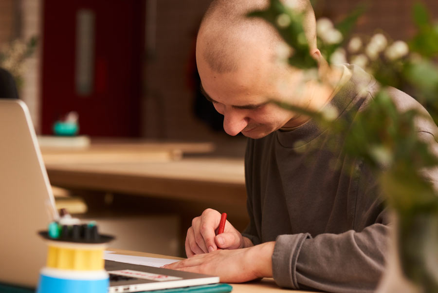 Man with a shaved head writing on paper at a desk with a laptop and electronic components nearby, partially obscured by green plant.