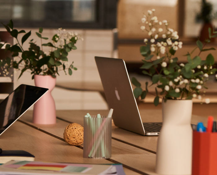 Modern workspace with laptops, potted plants in pastel vases, colored markers, and stationery on a wooden desk.
