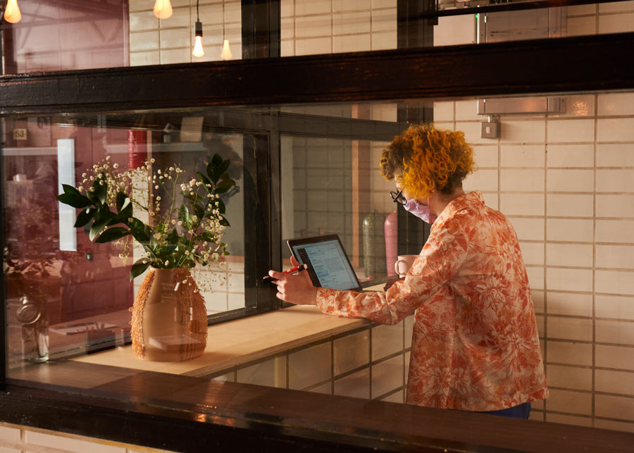 Person with curly orange hair and a floral shirt wearing a face mask, working on a tablet at a wooden counter next to a vase with green leaves and white flowers.