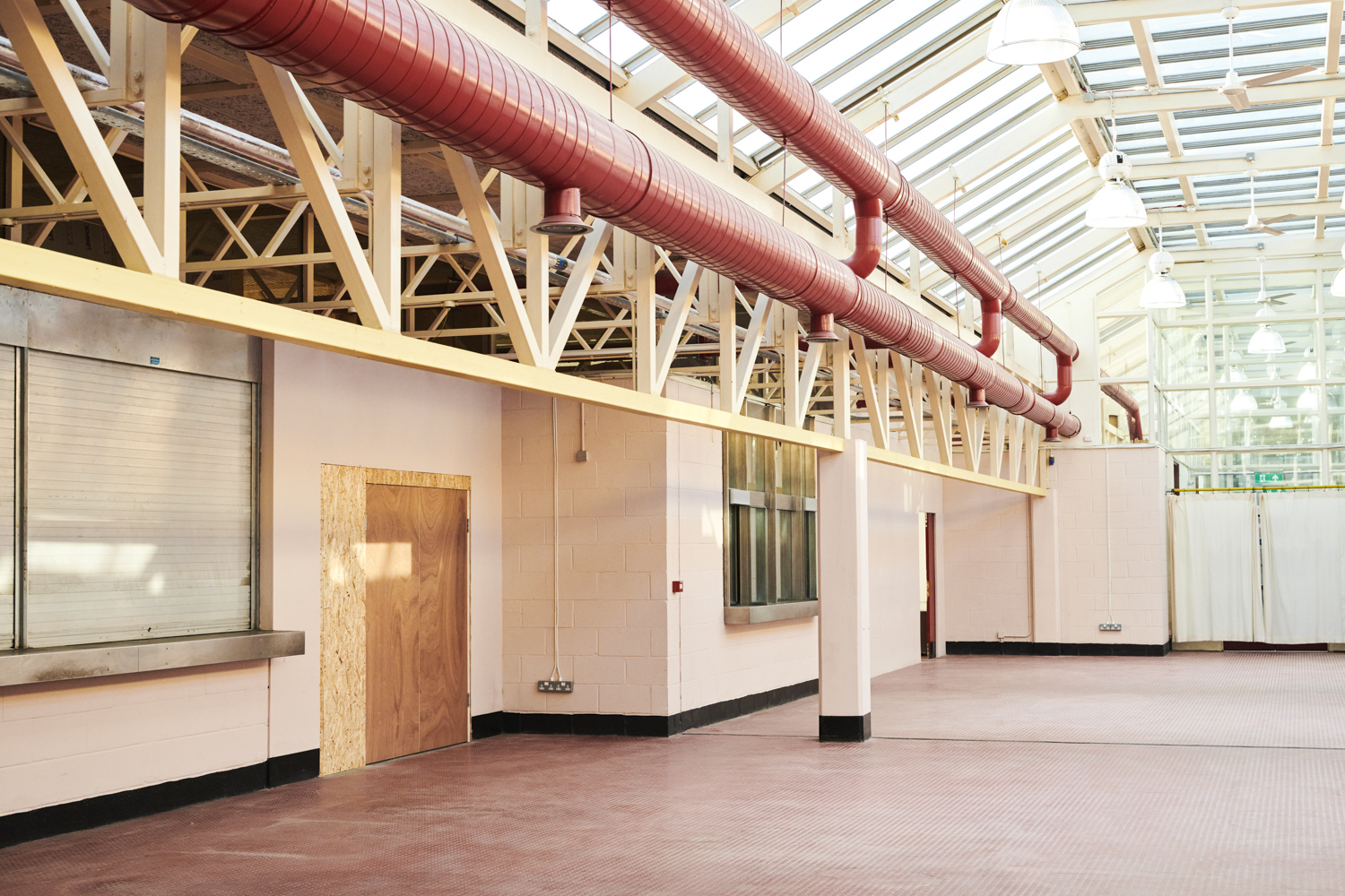 Bright industrial interior with large red ventilation pipes, pink walls, a closed shutter window, wooden door, and a glass ceiling with hanging lights.